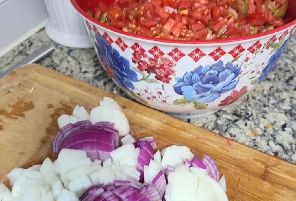 diced onions on a cutting board by a bowl of diced up tomatoes