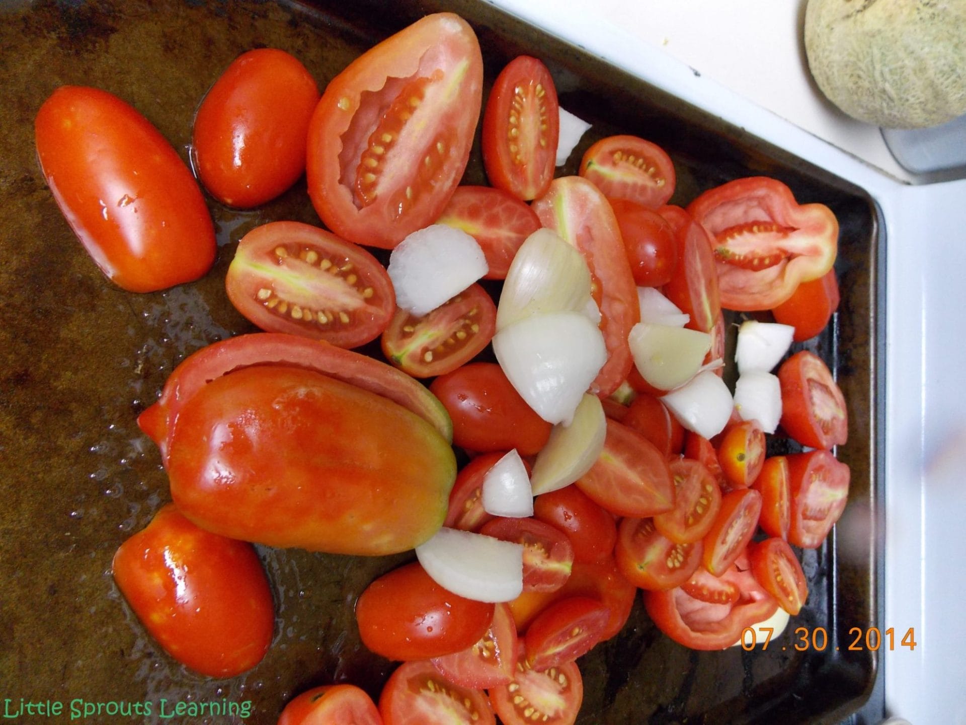 tomatoes cut in half on sheet pan with cloves of garlic and chopped onions for making roasted tomato sauce