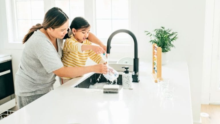 An adult and child teach manners as they wash dishes together at a kitchen sink on a bright day, with a plant and kitchen items softly framing the background.
