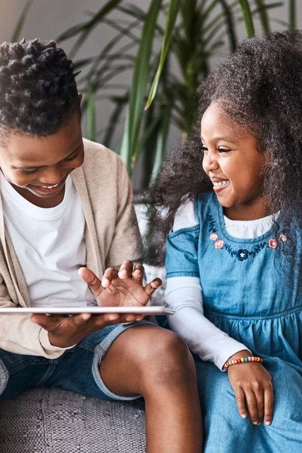Two children sit on a couch, smiling and looking at a tablet that subtly teaches manners. The boy wears a beige cardigan and shorts, while the girl dons a blue dress. A large plant is in the background.