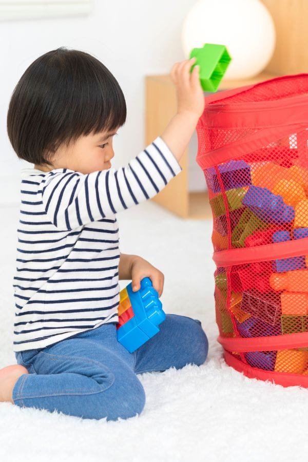 A child sits on a white carpet, playing with colorful plastic blocks next to a red mesh basket filled with more blocks, an environment that subtly begins to teach manners through sharing and cooperation.