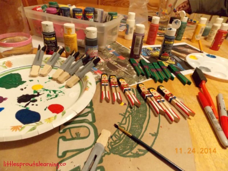 Table full of paints, brushes, markers, and plates of paint with clothespins to paint homemade superhero ornaments on.