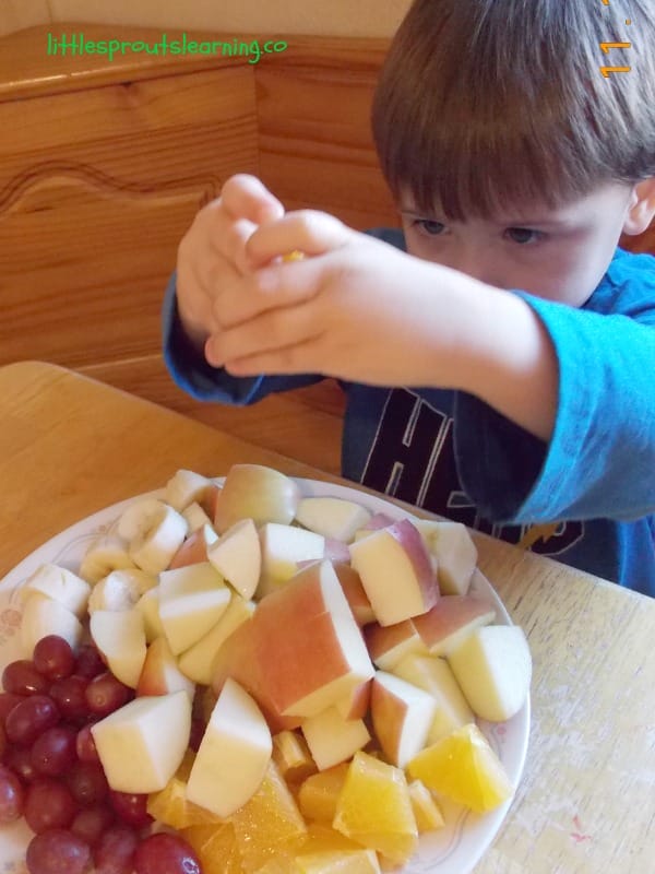 Child squeezing lemon juice on a plate of cut up fruit for fruit kabobs, bananas, oranges, grapes and apples.