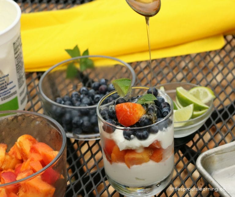 gorgeous fruit parfait healthy snack with yogurt and layers of peaches and blueberries on a table with lime wedges, blueberries, diced peaches, yogurt and a yellow napkin with a spoon above the parfait drizzling honey on