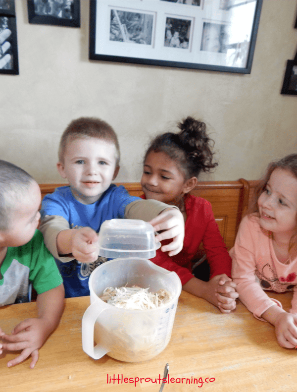 Four children sit at a table with a container of shredded material that resembles spaghetti pie. One child holds a lid above the container, while the others look engaged and curious.