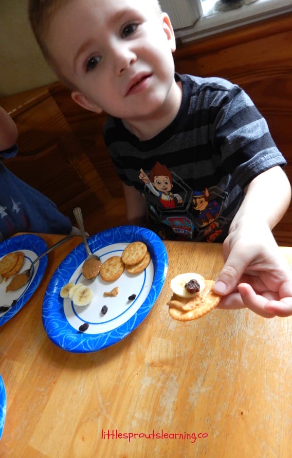 a child holding a ritz cracker fun snack that is spread with peanut butter and topped with a banana slice and a raisin to make a cat eye
