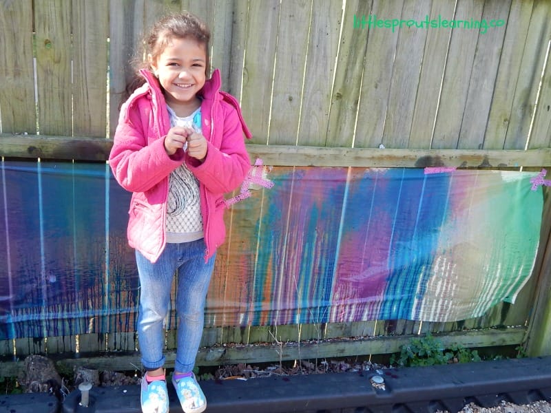 a child standing in front of a mural taped to a fence where children have sprayed as much paint as they wanted and let it drip down.