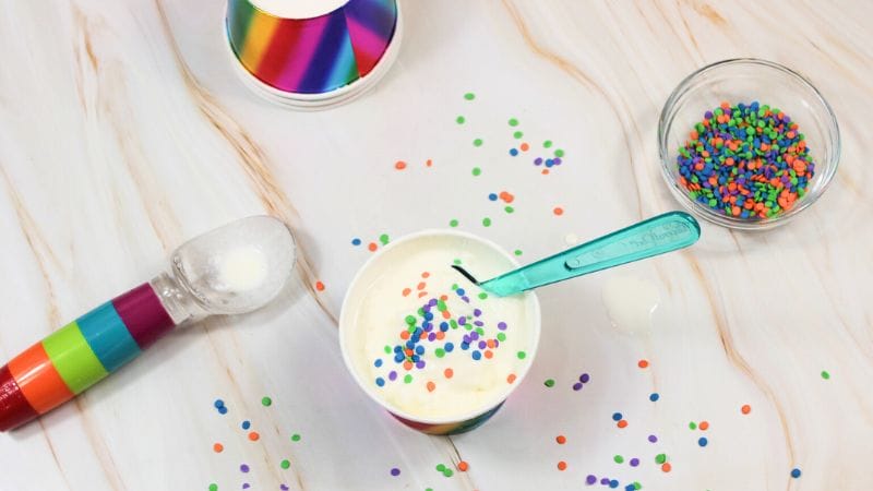 A cup of vanilla ice cream with colorful sprinkles, a turquoise spoon, a rainbow scoop, and a bowl of sprinkles on a marble surface—perfect for trying your favorite ice cream in a bag recipe at home.