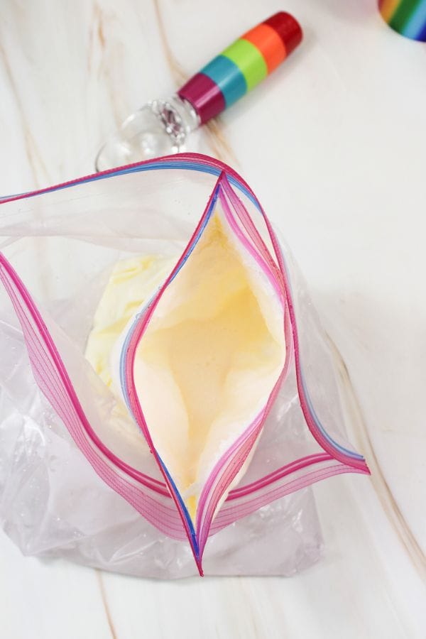 A plastic ziplock bag partially filled with yellow ice cream mixture sits on a white surface; a metal ice cream scoop with a rainbow handle is nearby, ready for this simple ice cream in a bag recipe.