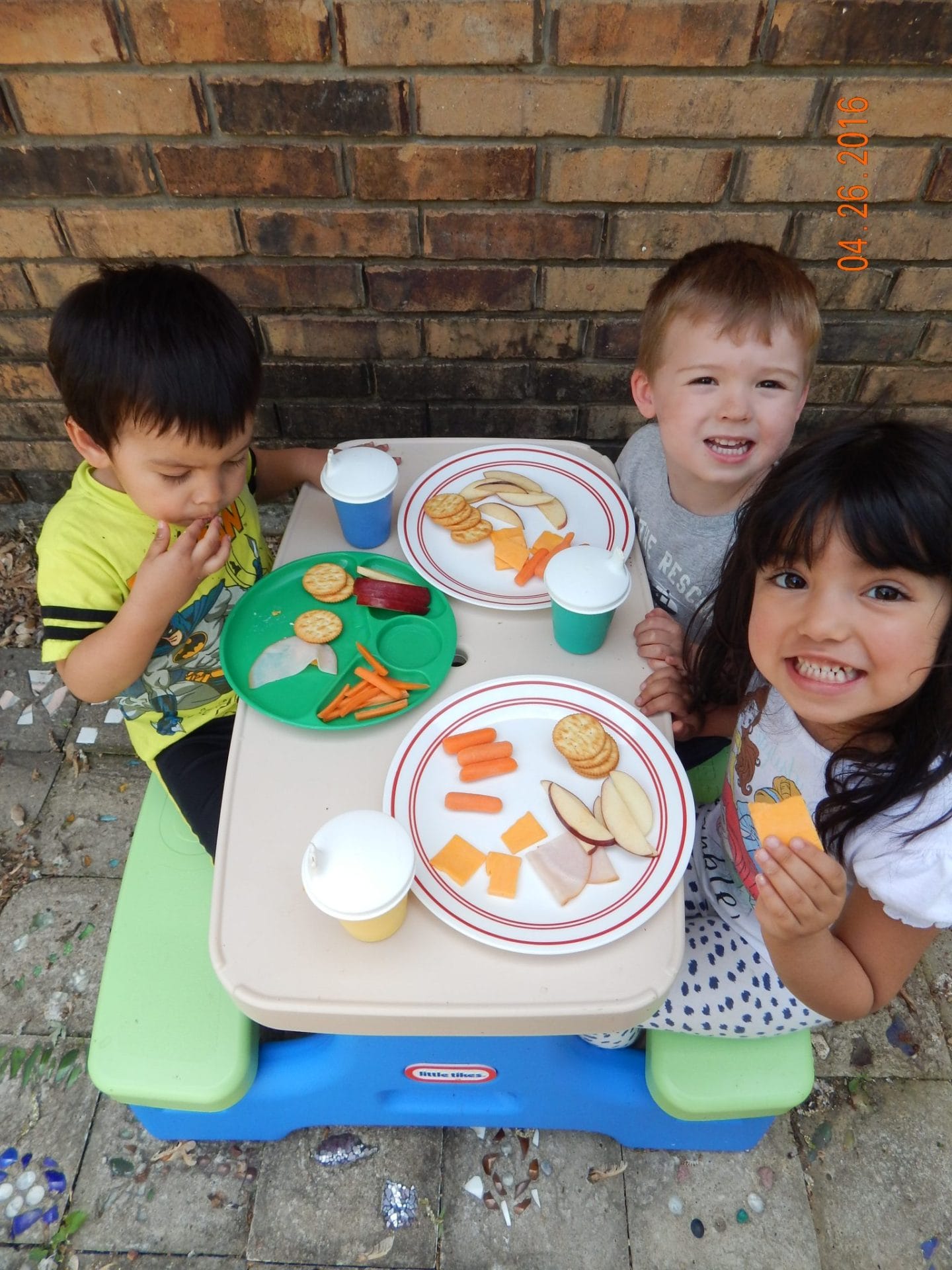 feeding toddlers healthy food for lunch, three kids sitting at a picnic table with plates of healthy food. They are eating and smiling. 