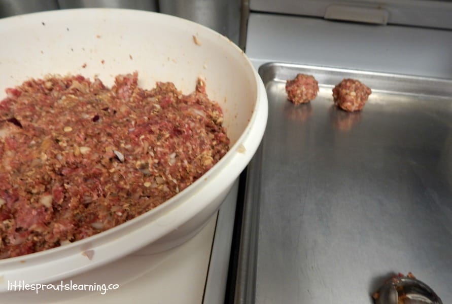 rolling out homemade meatballs for bulk cooking, a bowl of meatball mixture next to a sheet tray with two meatballs