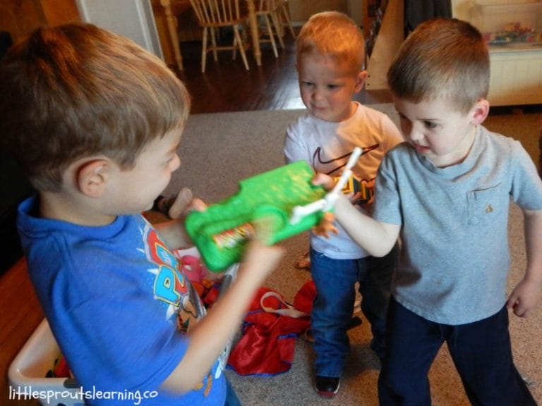 Three young boys play with a green toy, featuring an open mouth and teeth, in a living room setting.