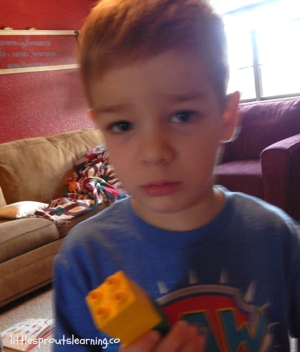 A child in a blue shirt, having a bad day, holds a yellow toy block in a living room with a sofa and a window in the background.