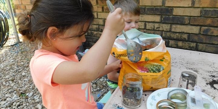 Kids making terrariums in the garden with a bag of dirt, some plants and mason jars for a garden activity for toddlers