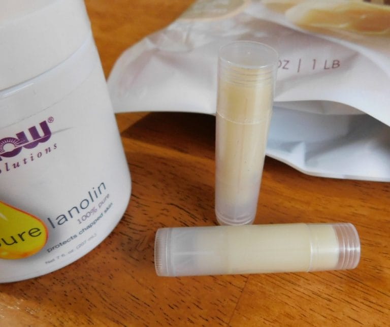 Two tubes of lip balm are next to an open container of pure lanolin lip balm and a sealed bag on a wooden surface.
