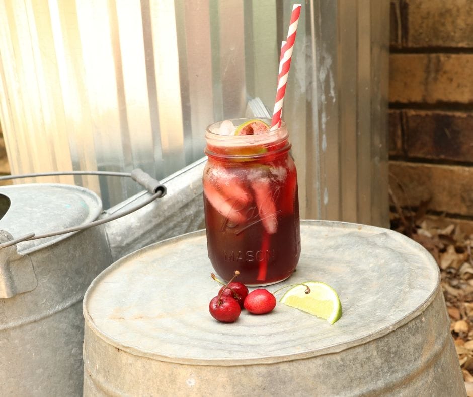 Cherry limeade in mason jar with paper straws and a lime wedge and three cherries on bucket in the garden.
