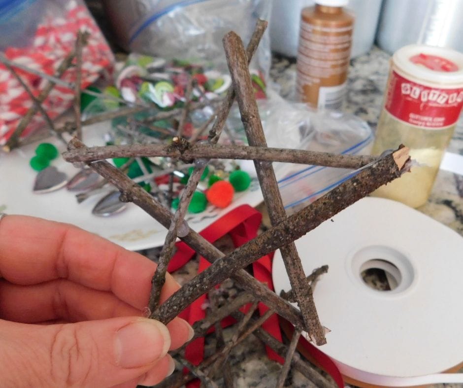 twigs glued together to make a star ornanament, held up in front of a table full of decorating supplies, glue, ribbon, pom poms and beads