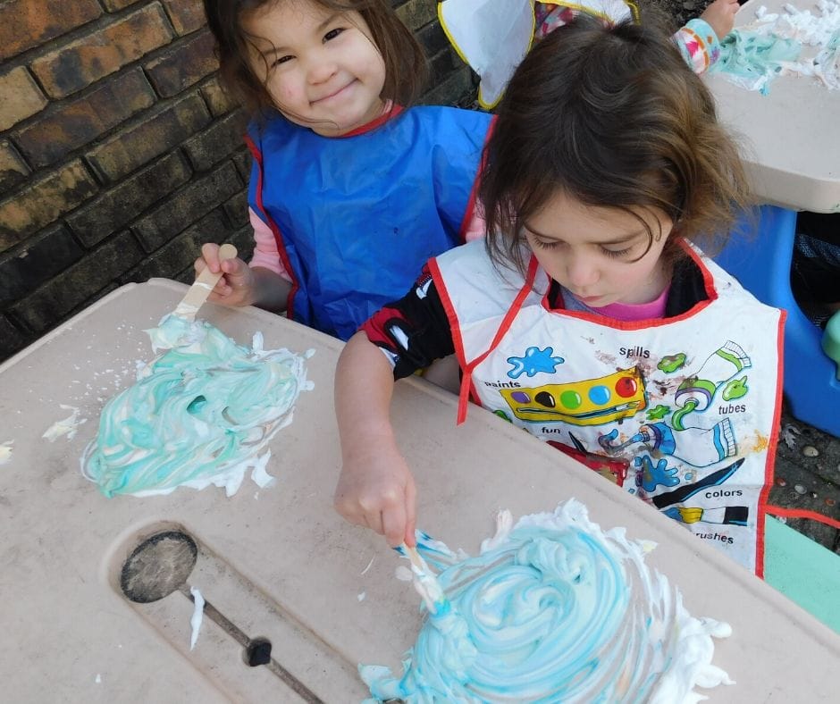 kids using shaving cream paint on a picnic table outside wearing smocks