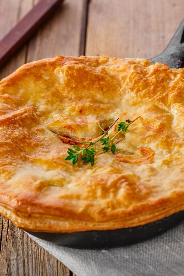 A golden-brown pork roast pot pie with a flaky crust, garnished with a sprig of fresh thyme, sits on a cloth atop a wooden table.