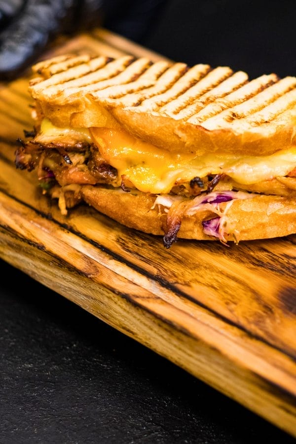Grilled panini sandwich with melted cheese, pulled pork roast, and slaw, served on a wooden board against a dark background.