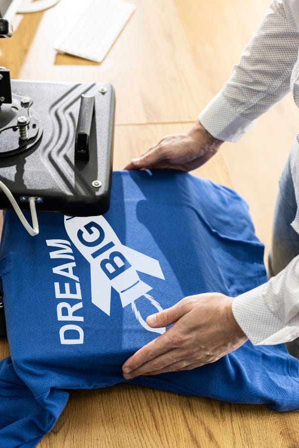 A person carefully places a blue "Dream Big" shirt under the heat press machine, ready to create inspiring child care T-shirts that spark imagination and hope.