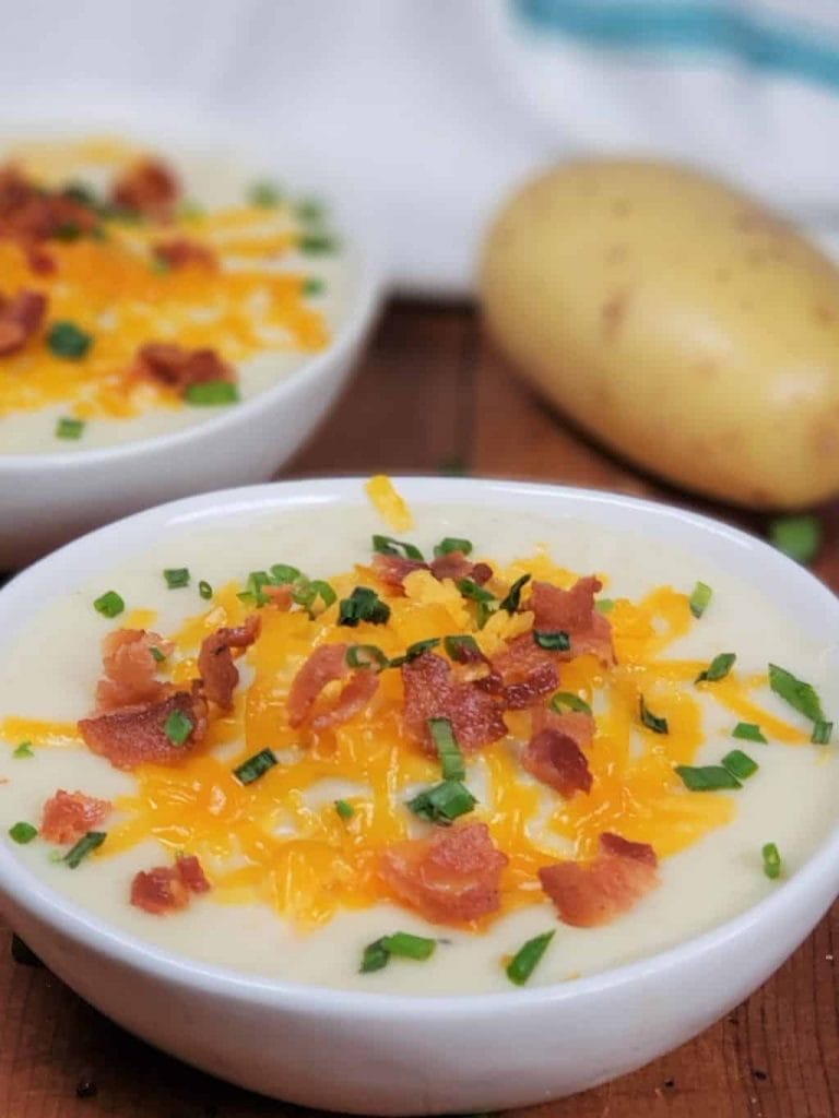 bowl of potato soup loaded with toppings on a cutting board with another bowl and a potato
