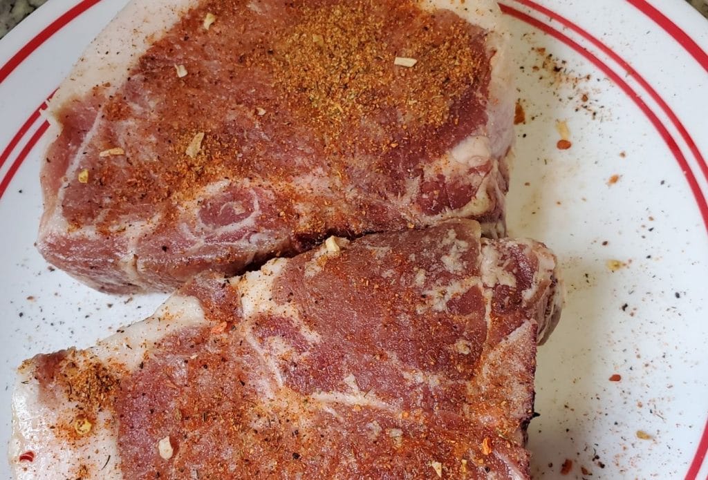 Two raw pork chops on a plate with seasoning.