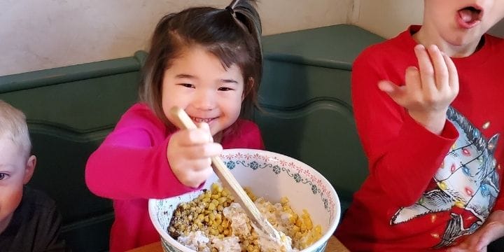 Kids sitting at a table taking turns adding ingredients to elote corn in a cup