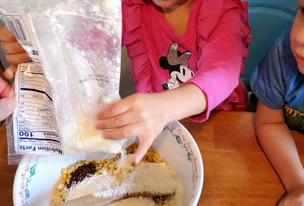 A child pouring cotija cheese into elote in a cup recipe