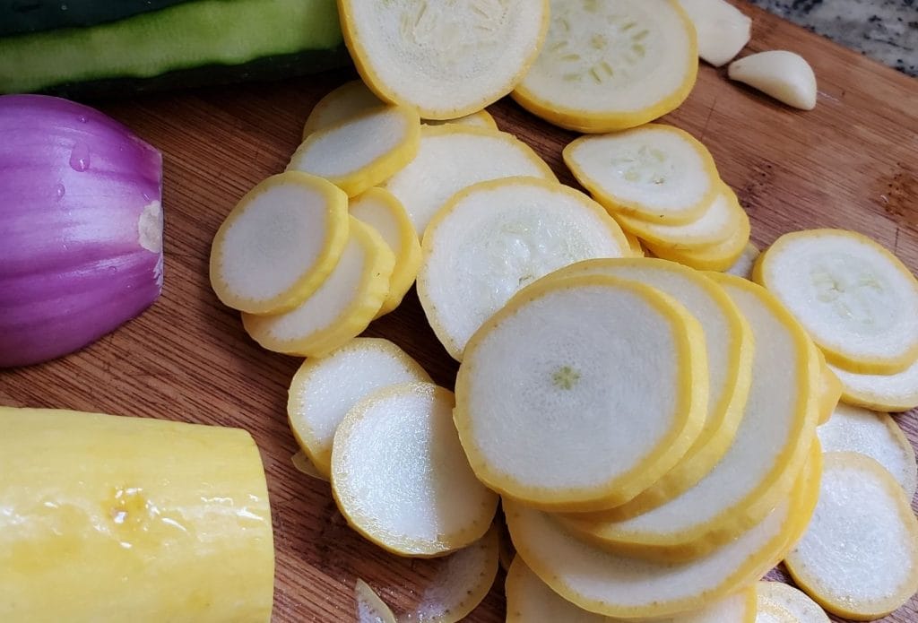 Yellow squash cut up on a cutting board next to whole squash, red onions, and cucumbers