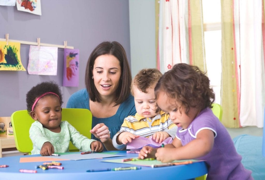 kids and a daycare teacher sitting at a table with crayons and paper