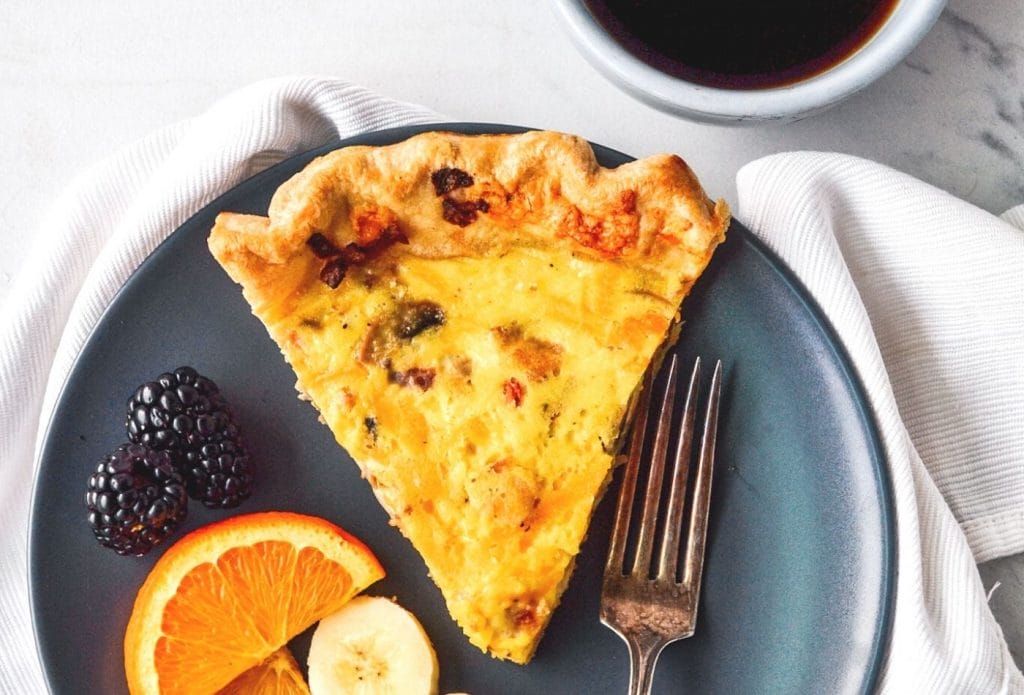 SLice of bacon and cheddar quiche on a plate with a fork and sliced fruit