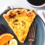 SLice of bacon and cheddar quiche on a plate with a fork and sliced fruit