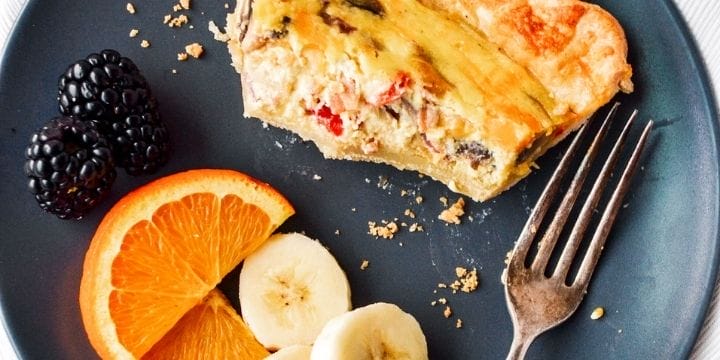 Bacon red pepper mushroom and cheddar quiche slice with bites gone on a plate with sliced fruit and a fork