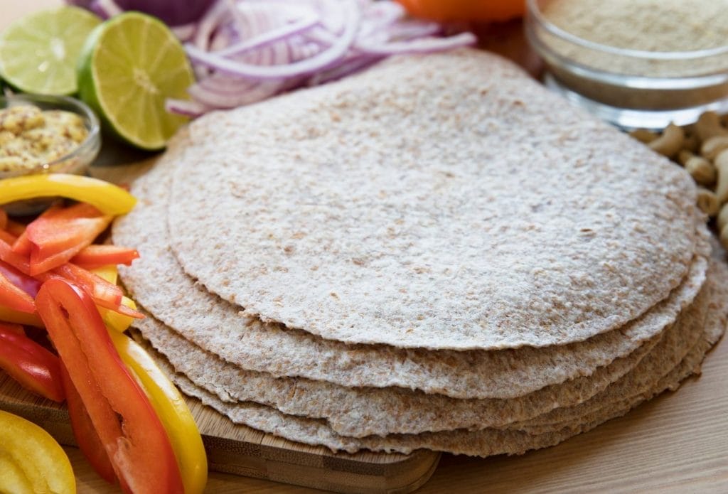 whole wheat tortillas in a pile on a board next to sliced vegetables to top bean burritos
