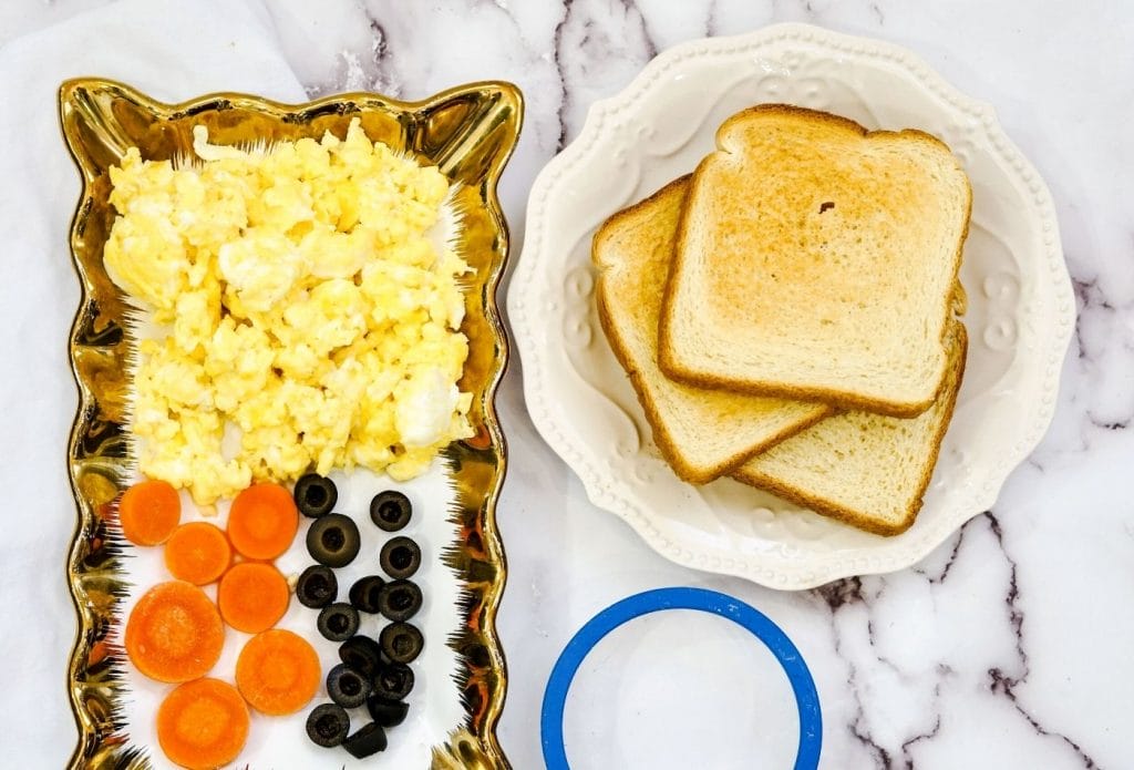 toast stacked on a plate next to a round cookie cutter and a tray with scrambled eggs and sliced carrots and black olives