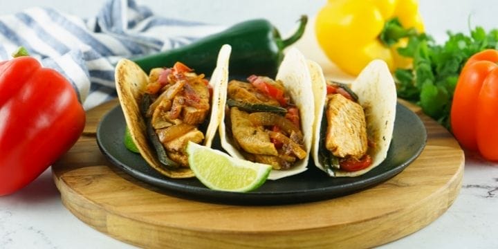 three chicken fajitas in tortillas on a plate with a wedge of lime and some fresh peppers and a towel