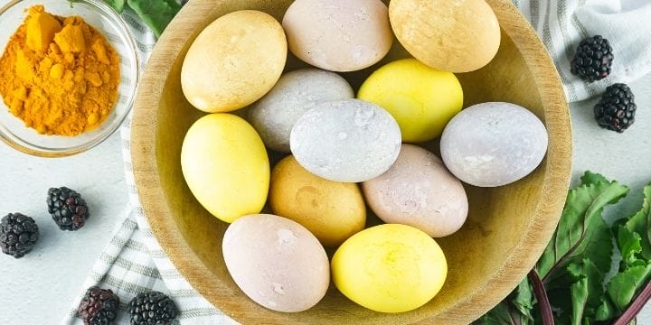 multicolored easter eggs dyed with natural dyes in a basket with blackberries, beet greens and turmeric on the table around the basket