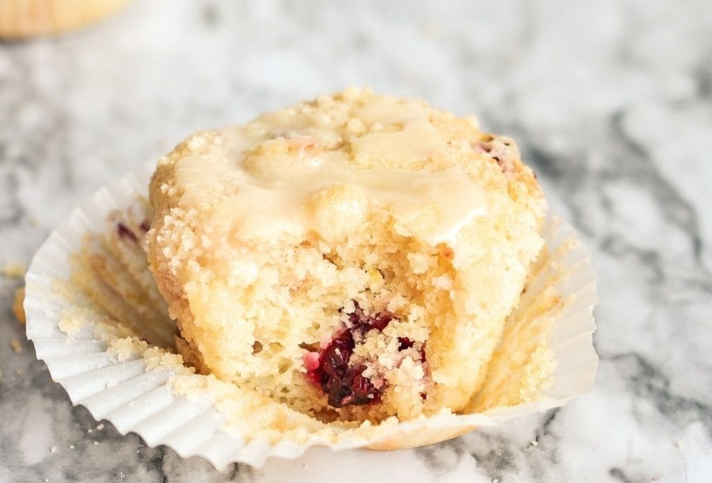 blackberry lemon crumb muffin with lemon drizzle in a cupcake paper peeled off and a bite out of it. The cooked blackberry is showing