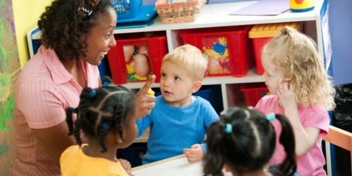 A woman engages with four young children seated at a table in a daycare classroom, colorful bins brightening the background.