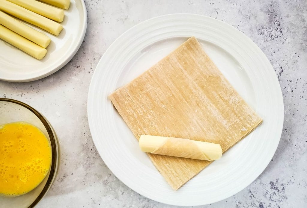 string cheese on a wonton wrapper next to a plate of cheese and a bowl of egg wash