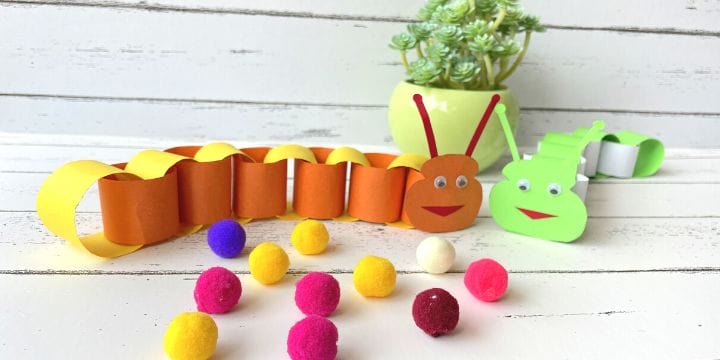 two paper caterpillars on a board with a plant and some colored pom poms