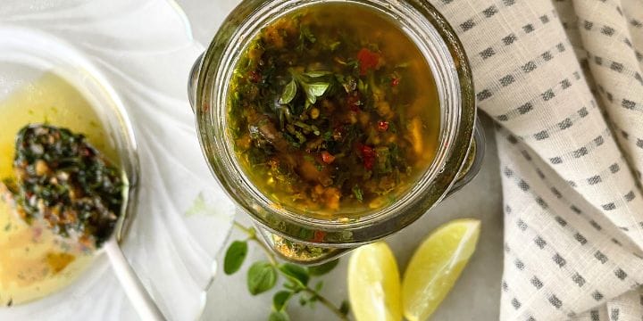 fresh garlic chimichurri sauce with fresh herbs in a jar next to fresh oregano and slices of lemon