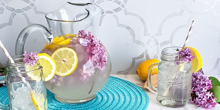 a pitcher of lilac lemonade on a mat next to a glass of lemonade
