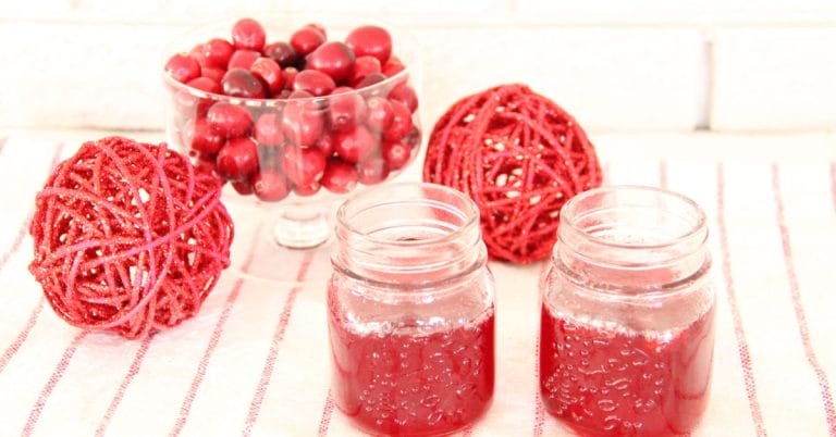 two jars of cranberry syrup on a board with balls and cranberries in a bowl