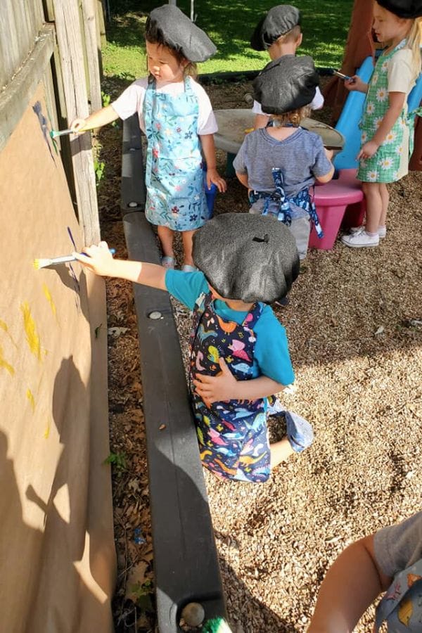 children painting on a large piece of paper with smocks and berets on