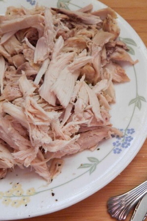 A plate of shredded cooked chicken, rests on a decorated white ceramic plate on a wooden table, with a fork partially visible.
