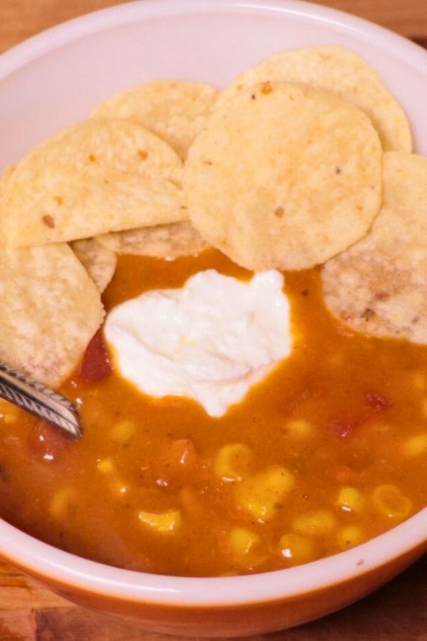 A bowl of soup with corn and tomatoes, topped with a dollop of sour cream, is surrounded by crispy tortilla chips.