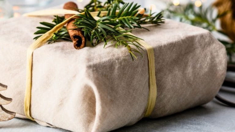 A fabric-wrapped gift is adorned with a cinnamon stick and evergreen sprigs, tied with a simple ribbon.