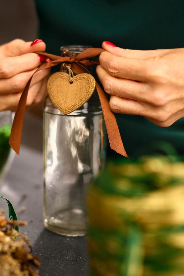 A person tying a brown ribbon with a heart-shaped tag around the neck of a clear glass jar,